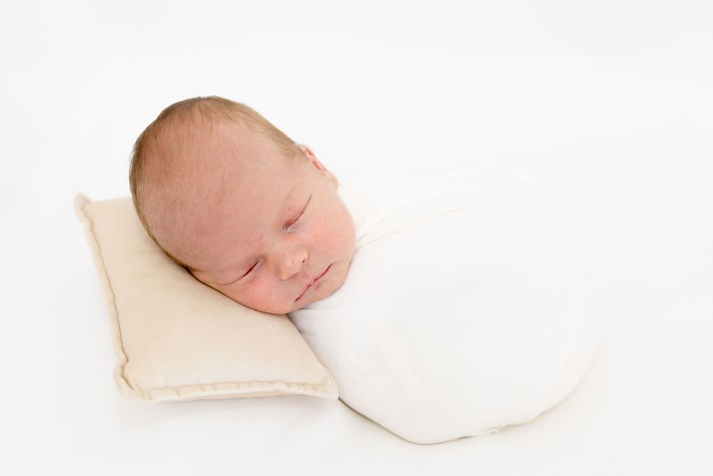 Peaceful newborn baby swaddled in white wrap sleeping on a small beige pillow against a white background.