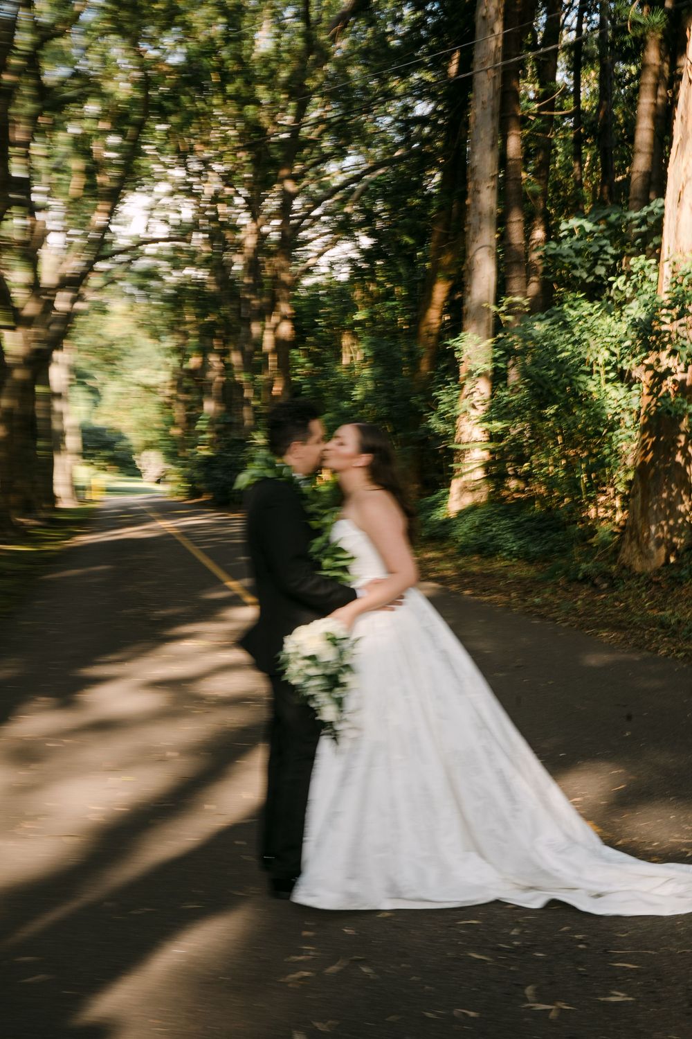 Bride and groom share a blurred romantic kiss on a tree-lined road, bride holding a bouquet.