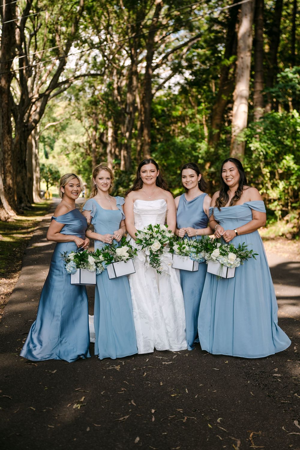Bride in white lace gown with four bridesmaids in dusty blue dresses holding white bouquets on a tree-lined path.