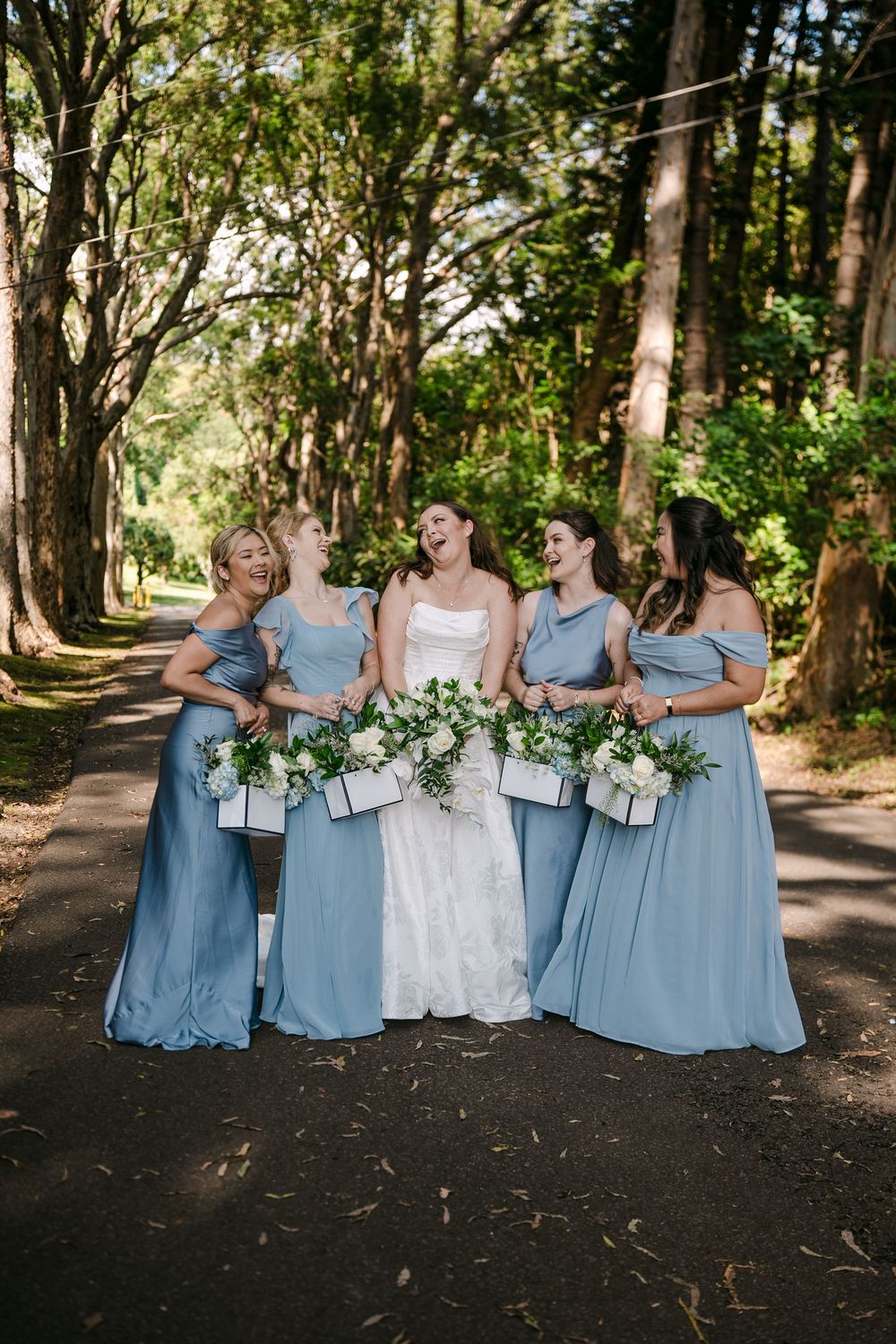 Bride in white lace gown laughs with four bridesmaids in dusty blue dresses holding white bouquets on a tree-lined path.