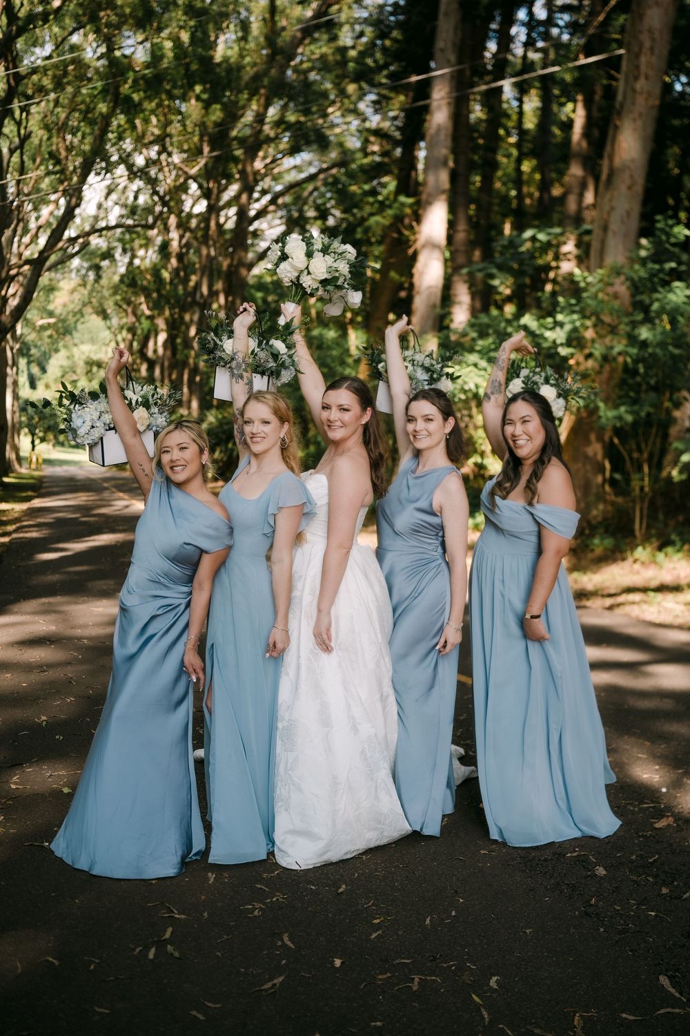 Bride in white gown poses with four bridesmaids in dusty blue dresses on a tree-lined path.