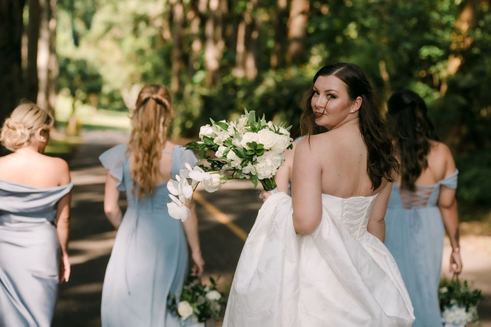 Bride in white strapless gown looks back smiling holding white bouquet with bridesmaids in blue dresses on tree-lined path.