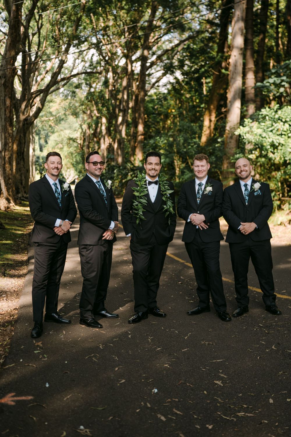 Groom and groomsmen in dark suits pose on a tree-lined path at an outdoor wedding ceremony.