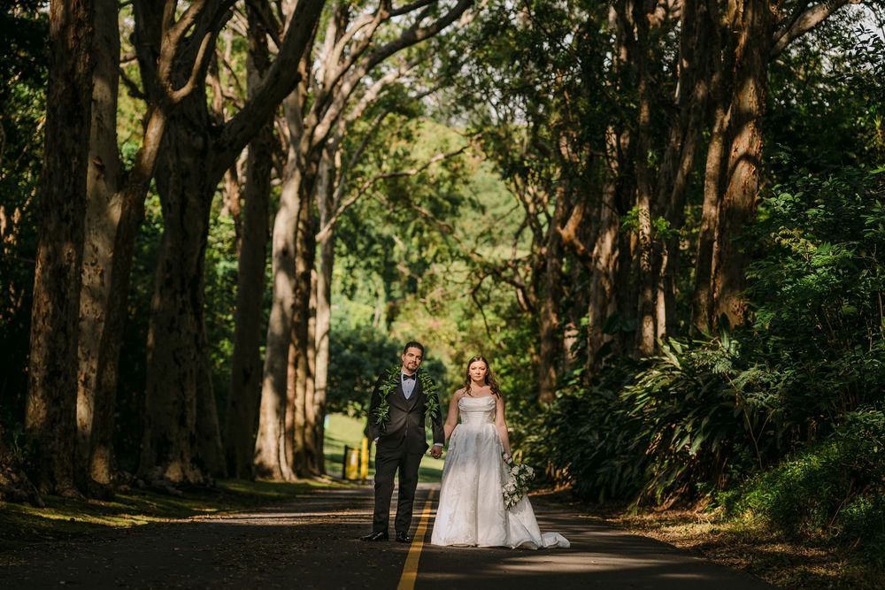 Bride and groom walk hand-in-hand down a tree-lined road surrounded by lush green foliage.