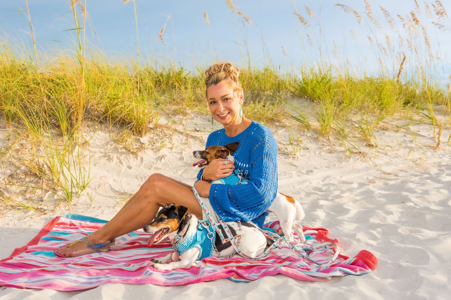 Woman in blue dress sits on beach blanket holding small dog, smiling with sand dunes and sea oats behind her.