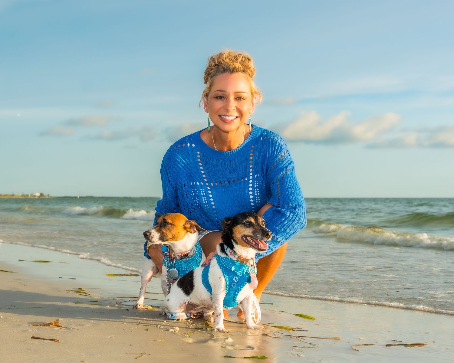Woman in blue sweater kneeling on beach with two Jack Russell Terriers wearing matching blue outfits.