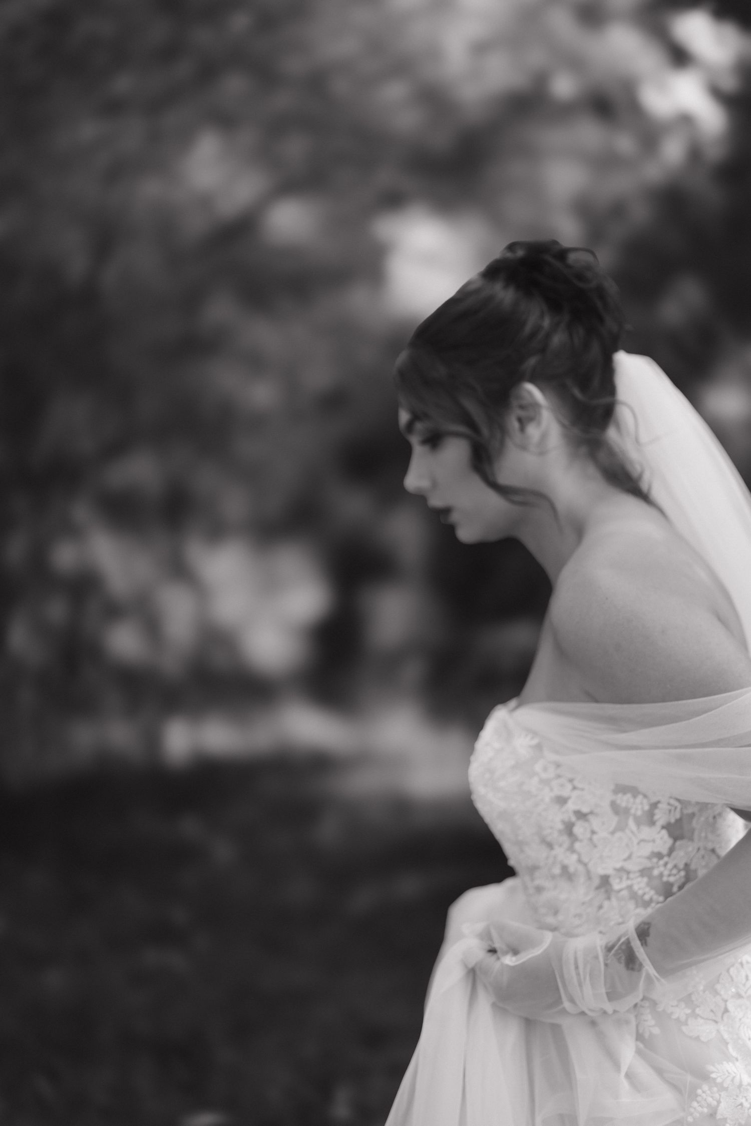 Black and white profile portrait of a bride in lace gown and veil, looking down in an outdoor setting.
