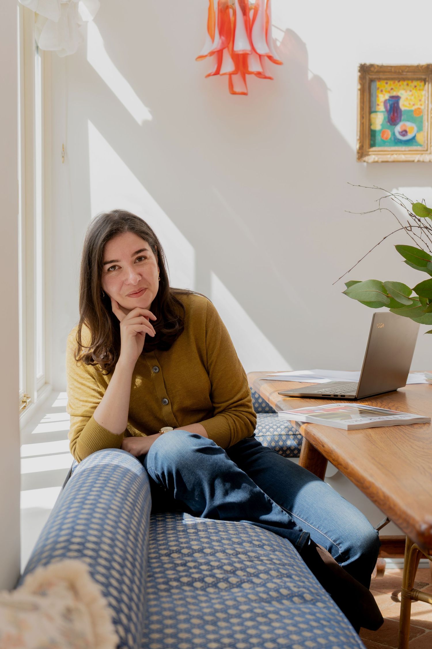 A smiling woman in a green shirt and jeans sits barefoot on a wooden stool in a bright, plant-filled kitchen.