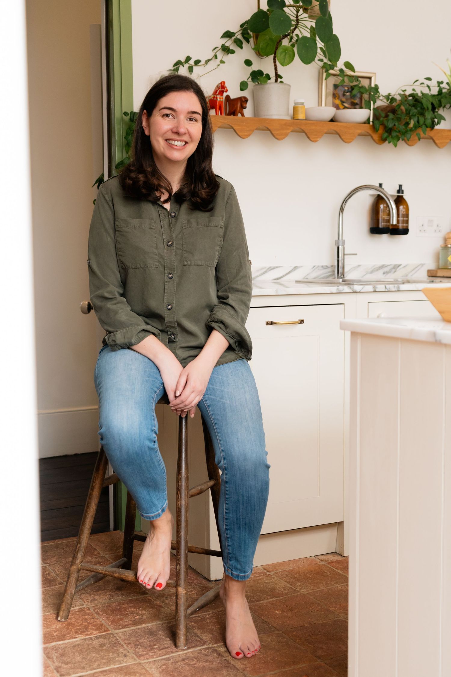 A smiling woman in a green shirt and jeans sits barefoot on a wooden stool in a bright, plant-filled kitchen.