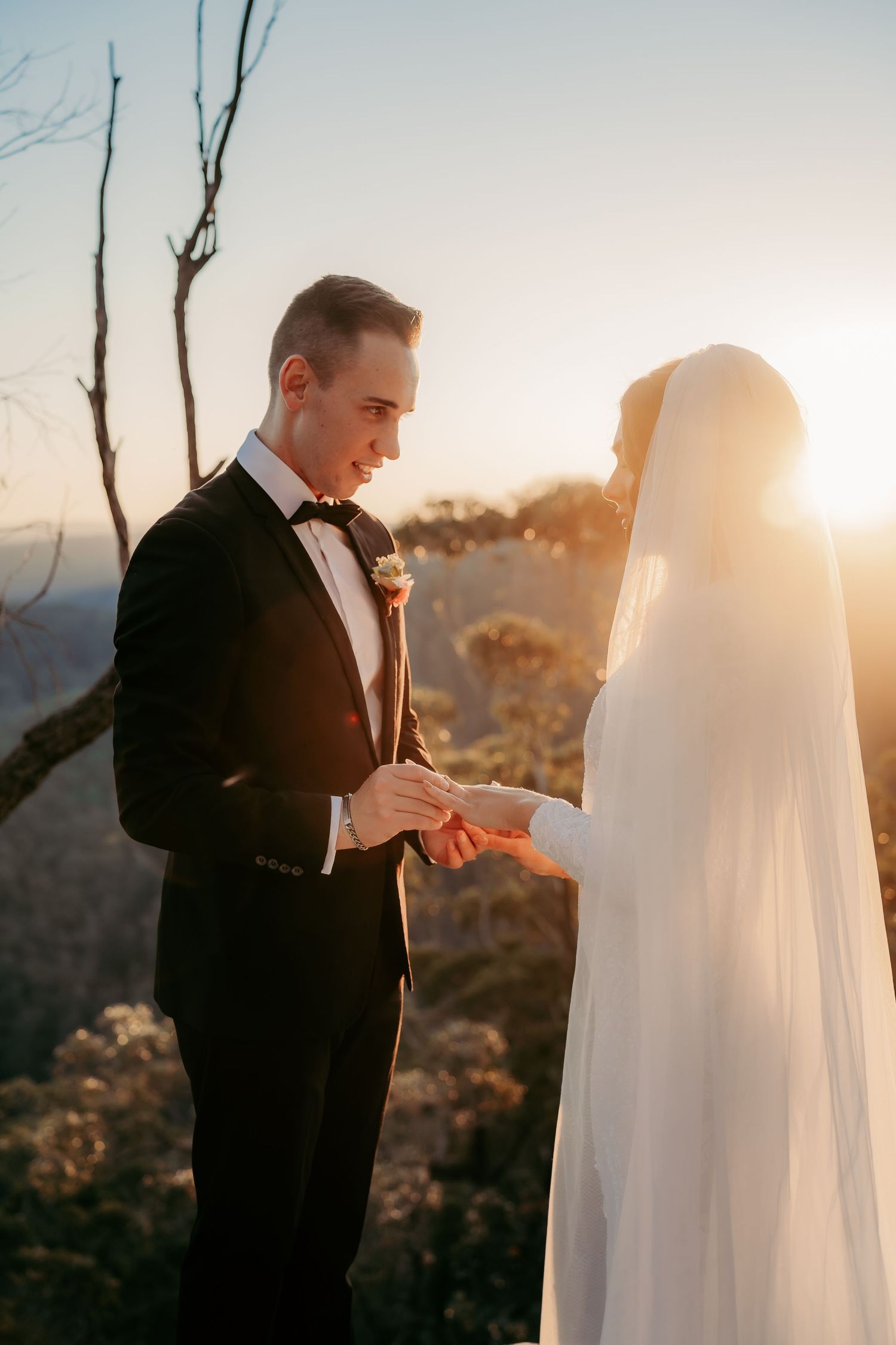 Groom placing wedding ring on bride's finger at golden hour sunset with mountain backdrop.