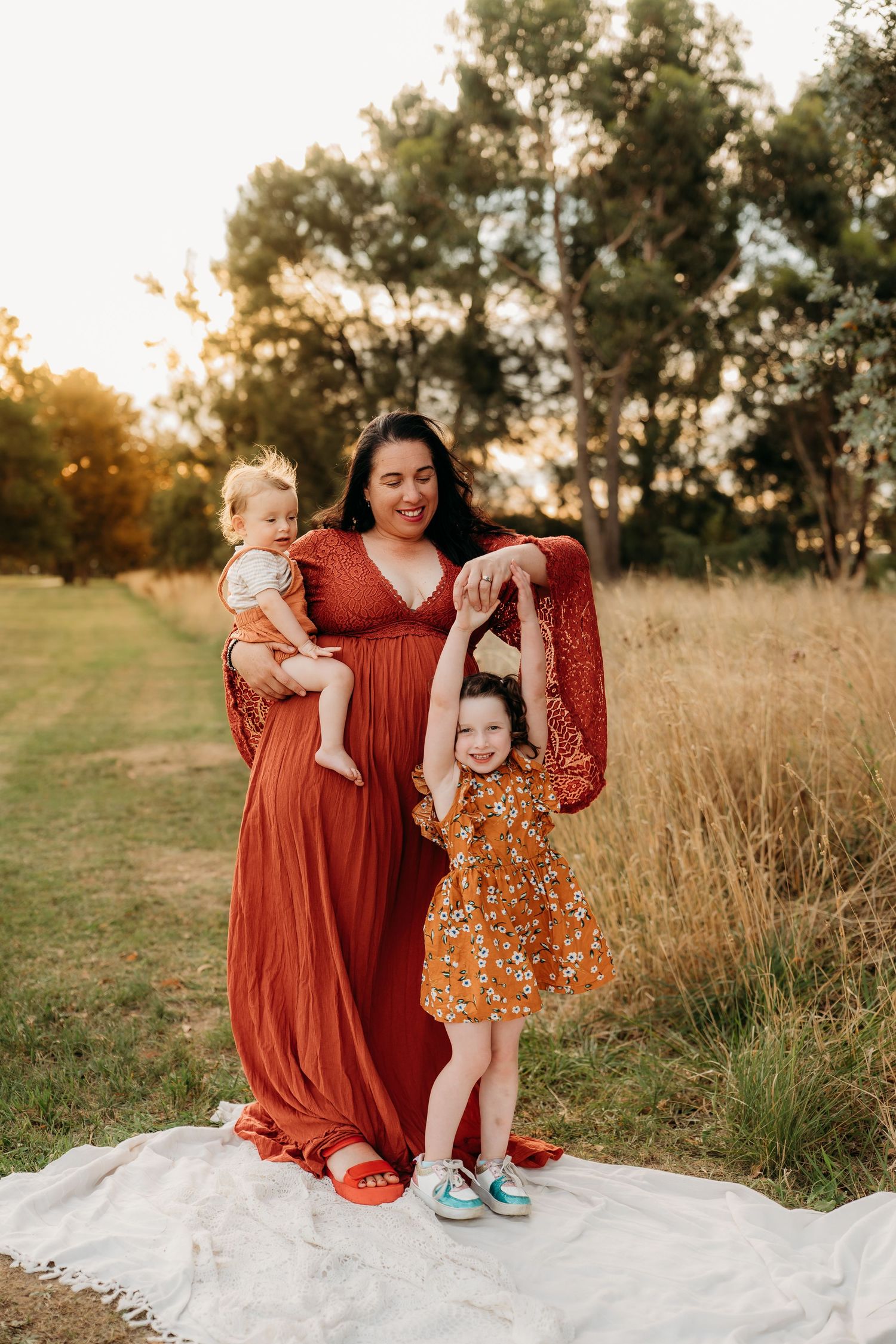 A mother in a rust red maxi dress holds her baby while her toddler daughter dances in a sunny outdoor field.