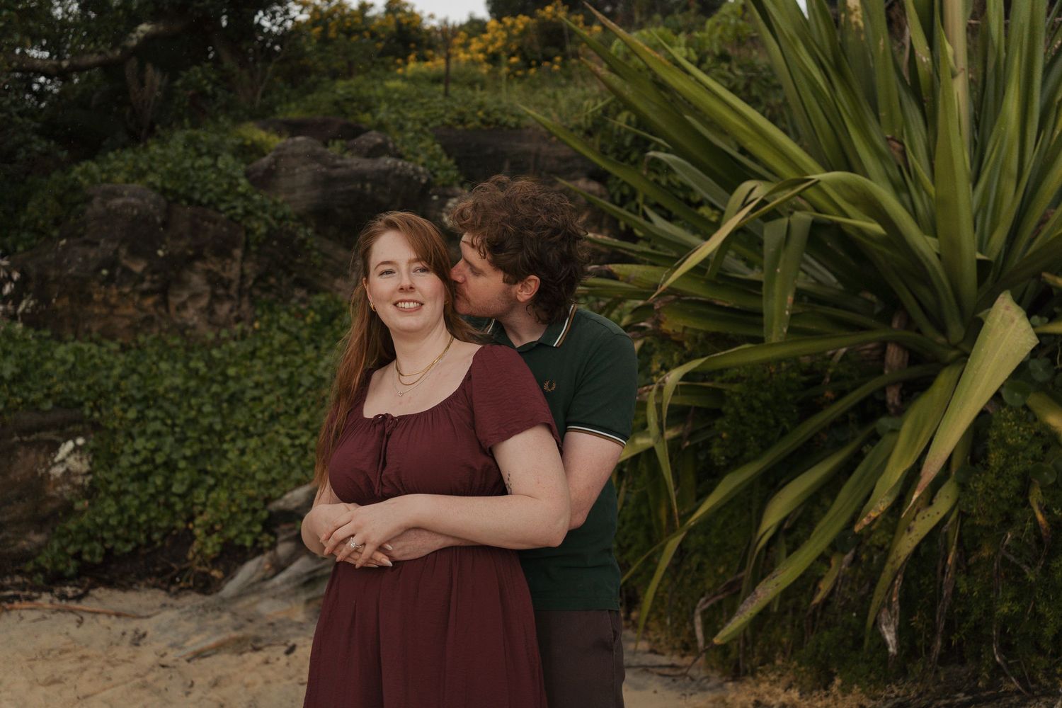 Couple sharing a tender moment on a sandy beach, woman in burgundy dress, lush tropical plants in background during proposal photoshoot sydn