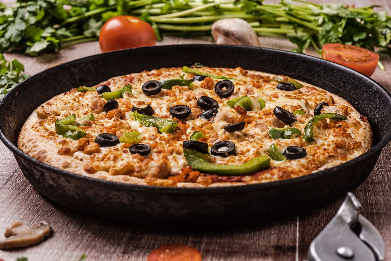 Cast iron pan pizza topped with black olives, green peppers, and melted cheese on a rustic wooden table.