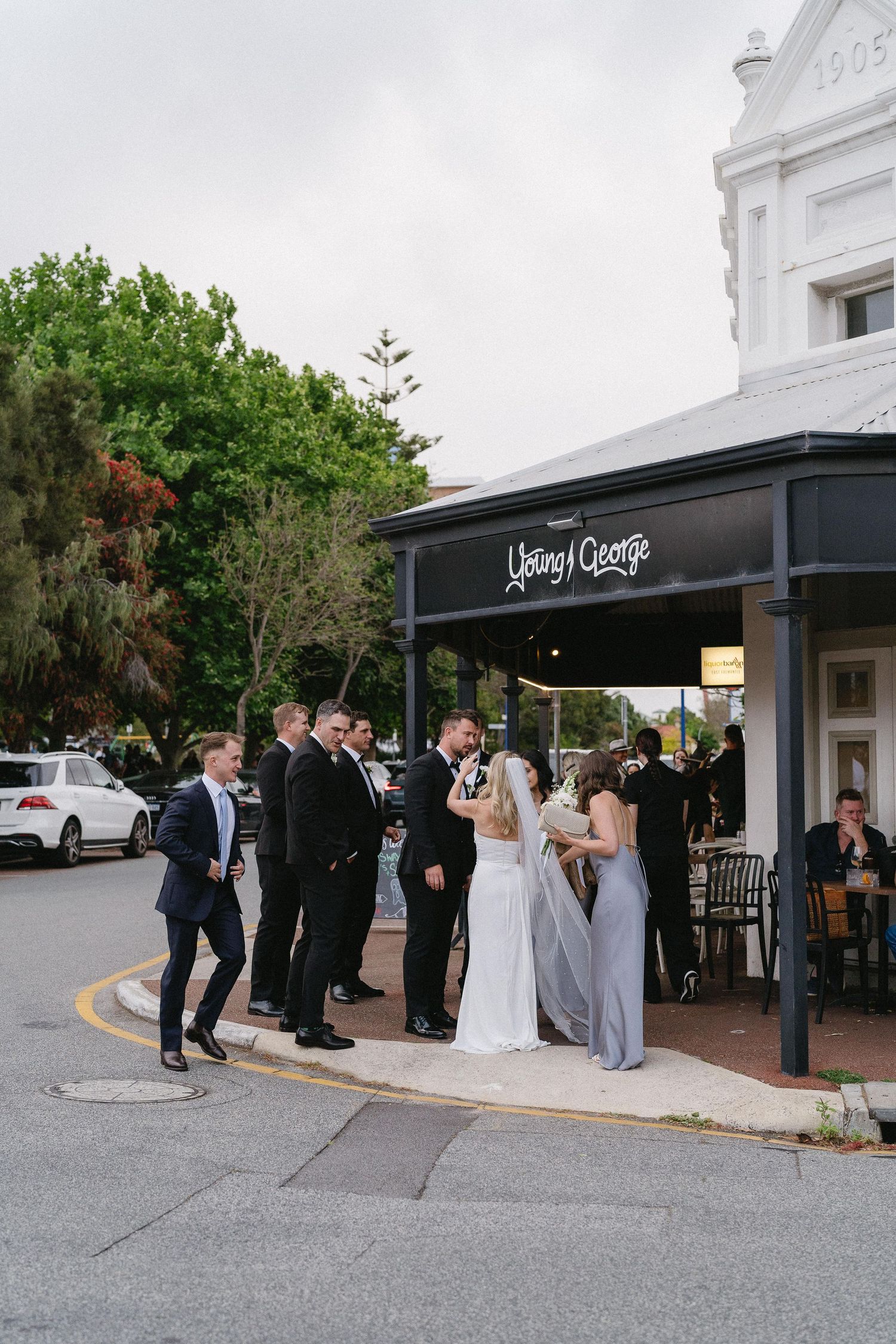 Bride in white gown laughing joyfully at wedding reception with two men in tuxedos behind her.