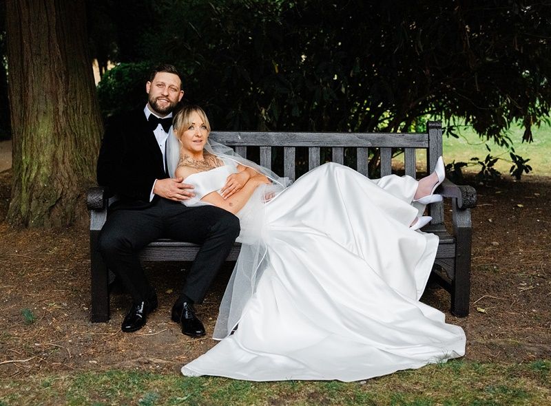 Bride in white gown reclines on groom's lap on a wooden bench under a tree in an elegant outdoor wedding portrait.