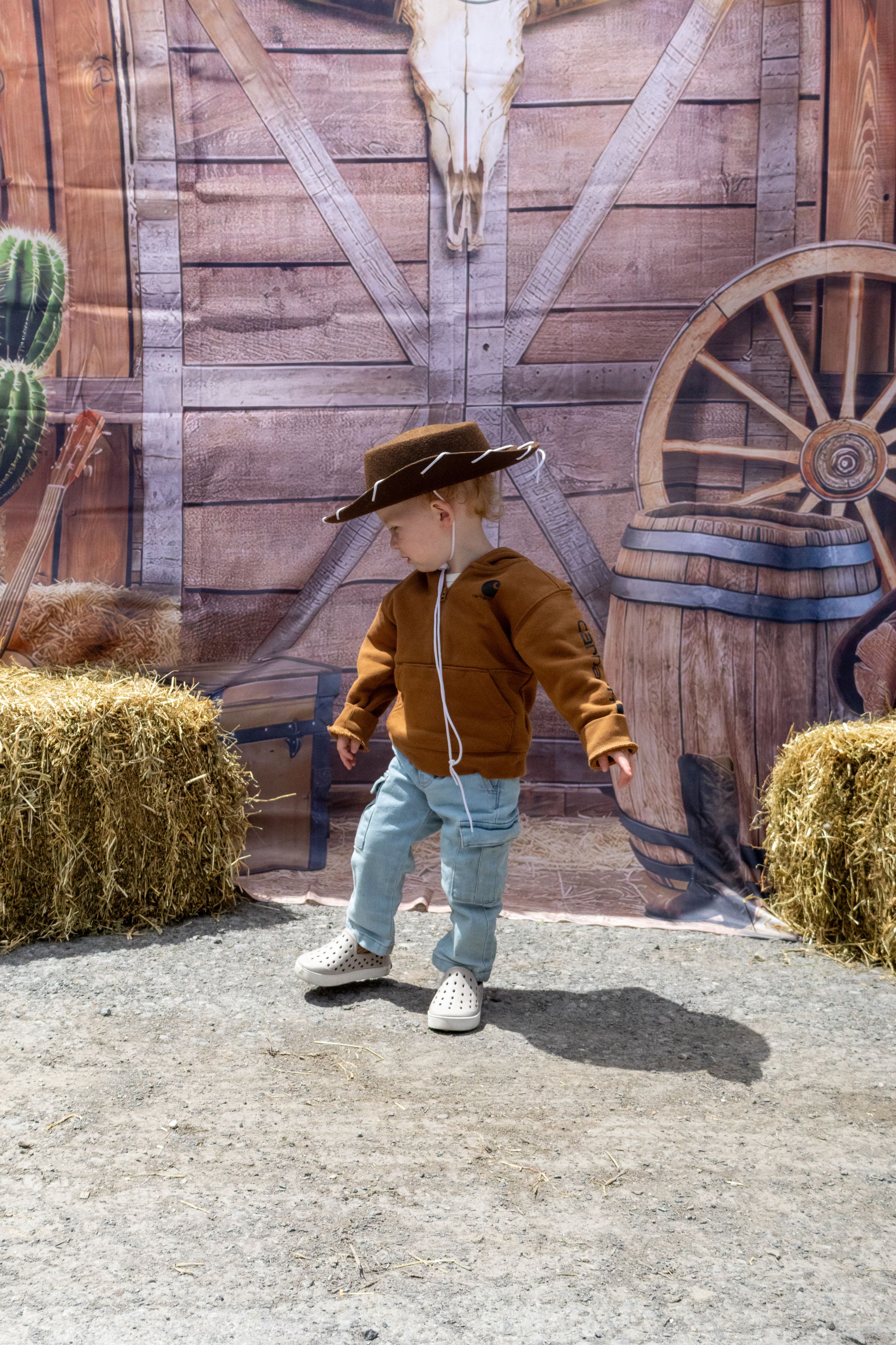 Toddler dressed as a cowboy in brown jacket, jeans, and oversized hat poses in front of Western-themed backdrop.