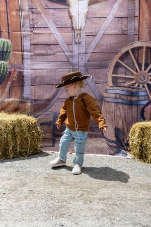 Toddler dressed as a cowboy in brown jacket, jeans, and oversized hat poses in front of Western-themed backdrop.