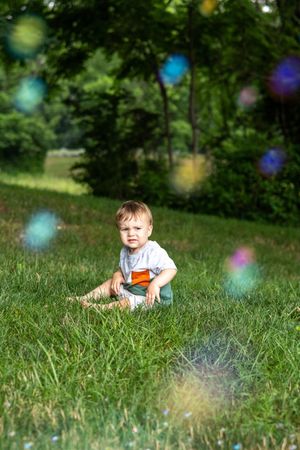 Toddler boy sitting on green grass in a sunny park surrounded by colorful soap bubbles floating in the air.
