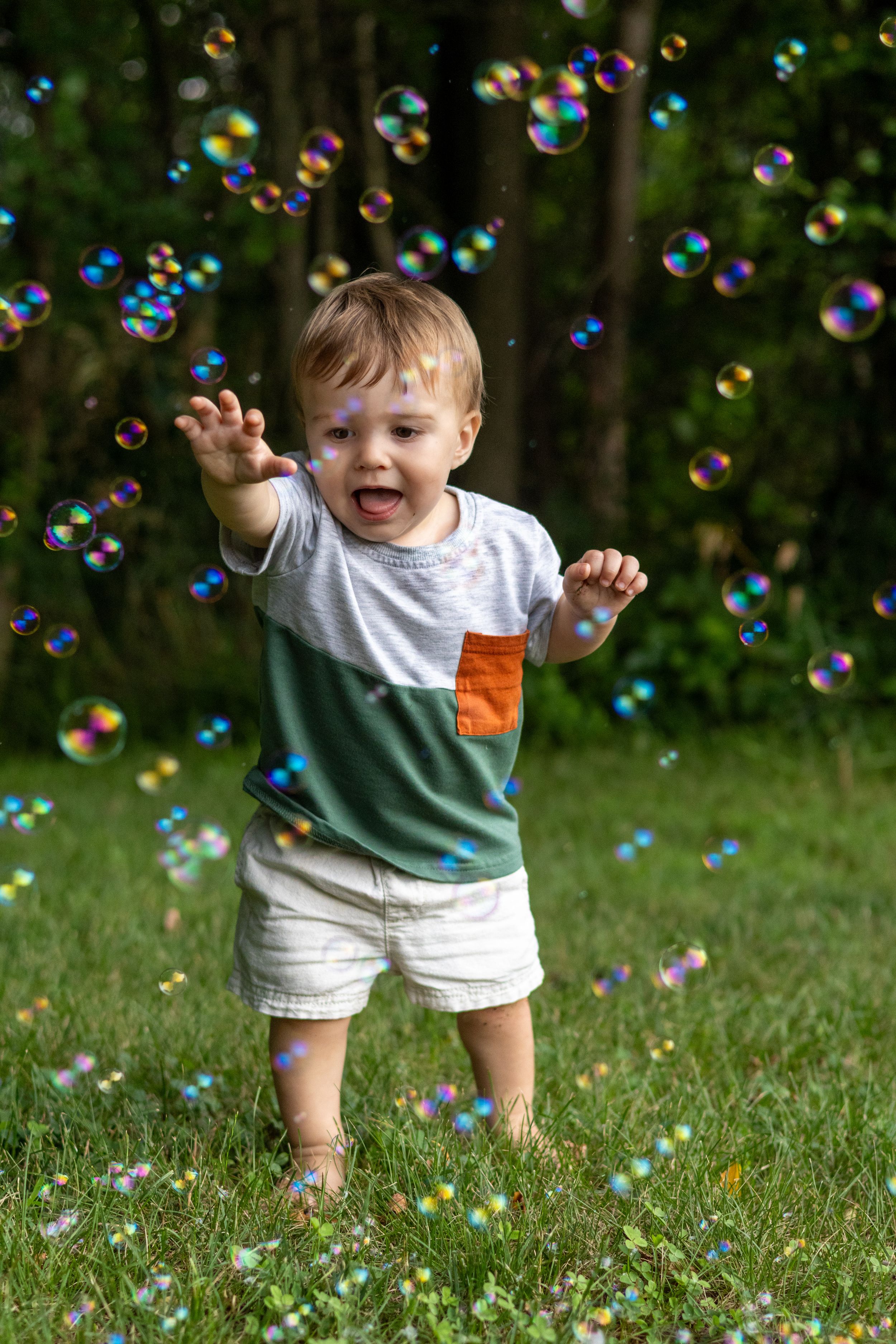Joyful toddler boy reaching for colorful soap bubbles floating in a sunny green backyard garden.