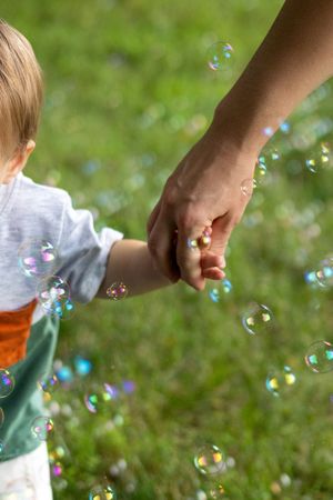 Adult holding a toddler's hand outdoors on green grass surrounded by colorful floating soap bubbles.