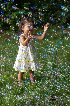 A joyful toddler girl in a white floral dress plays with colorful soap bubbles on a sunny green lawn.