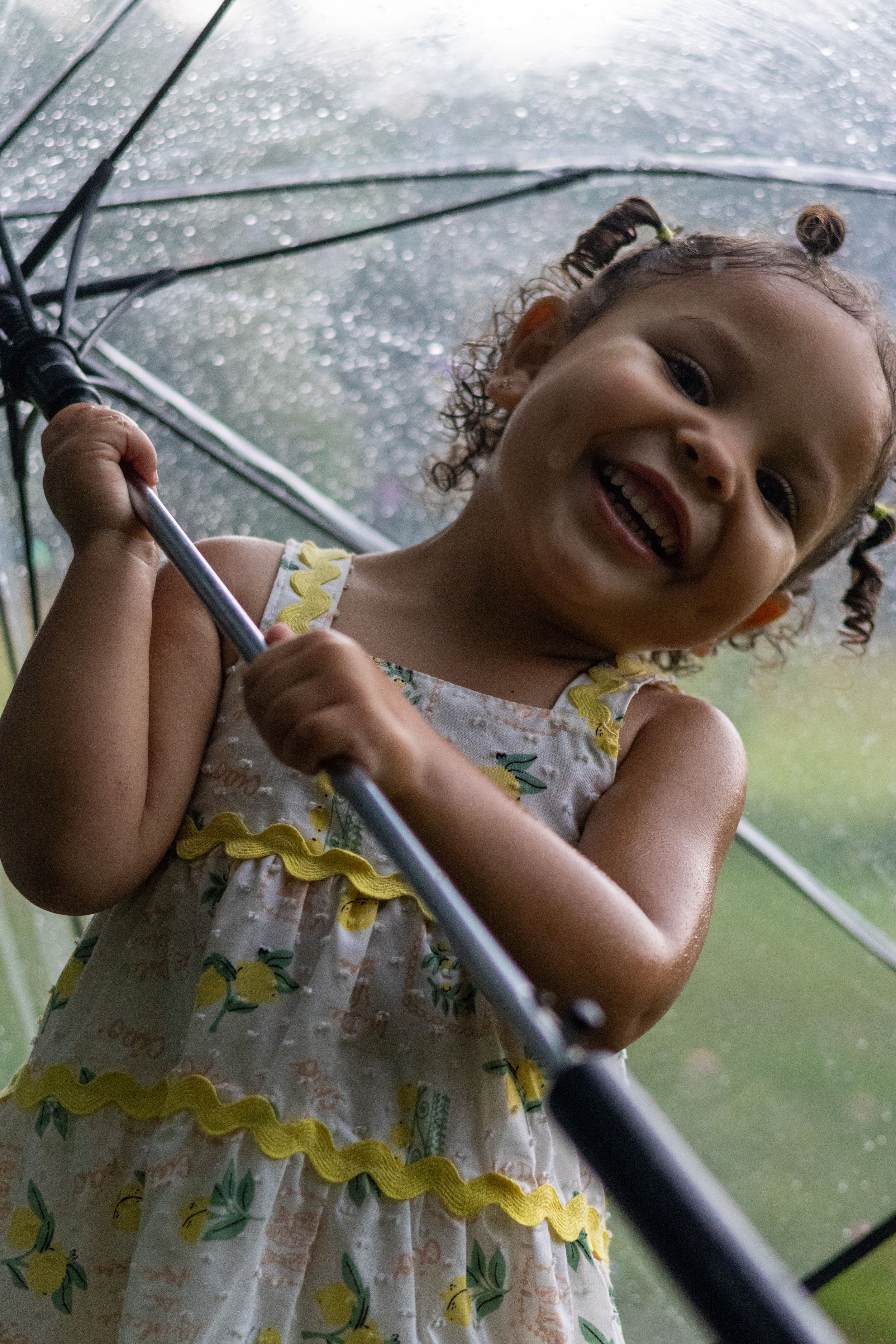 Smiling toddler girl in a floral yellow dress holds a clear umbrella in the rain, looking joyful outdoors.