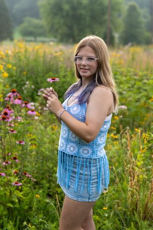 Young blonde teen girl in blue crochet top holding a pink flower in a colorful wildflower meadow.