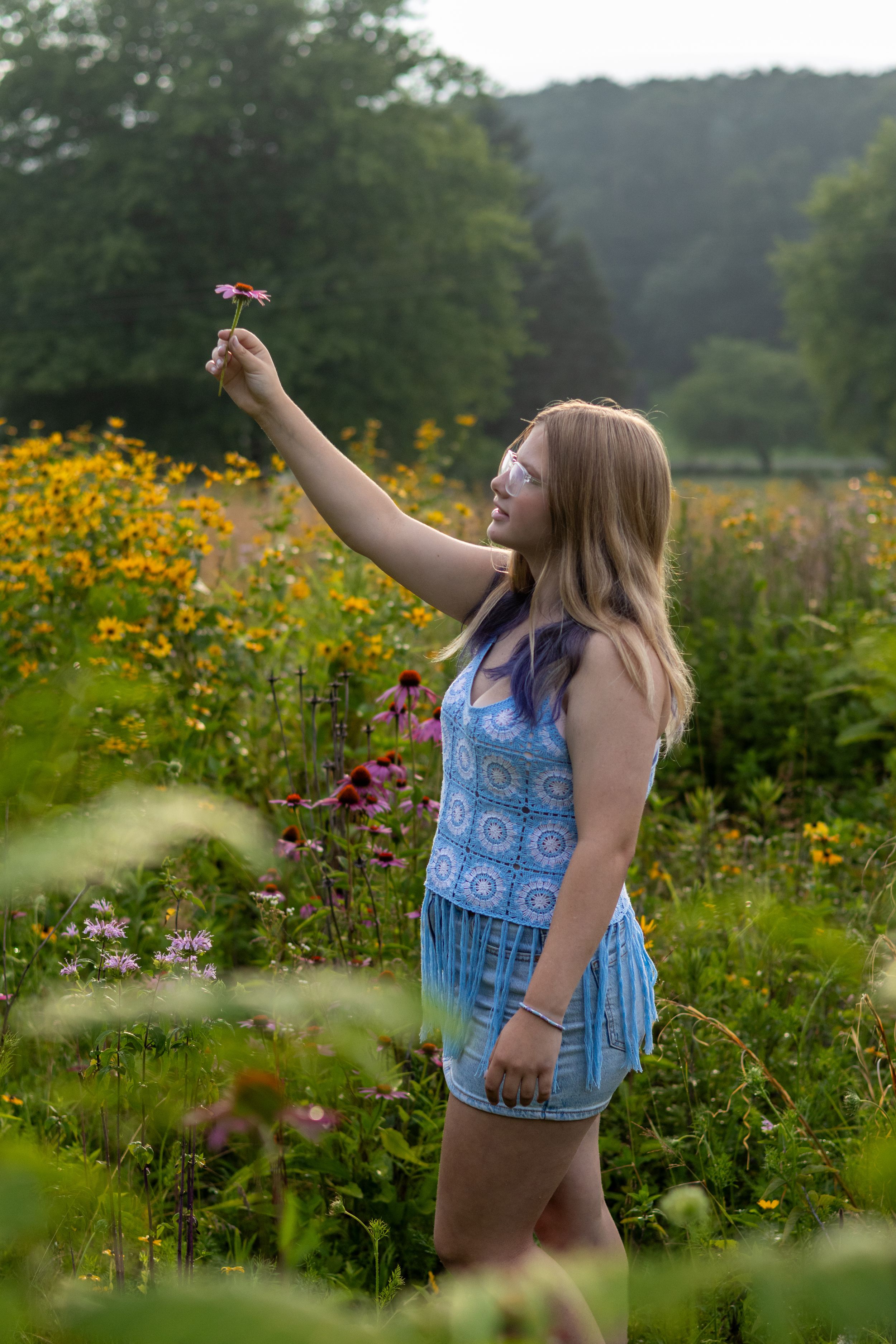 Young woman in blue romper holding a wildflower in a colorful meadow garden with yellow and pink blooms.