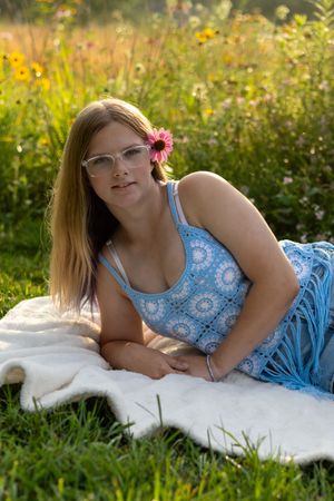 Young woman with glasses and pink flower in hair lying on white blanket in sunny wildflower meadow wearing blue crochet dress.