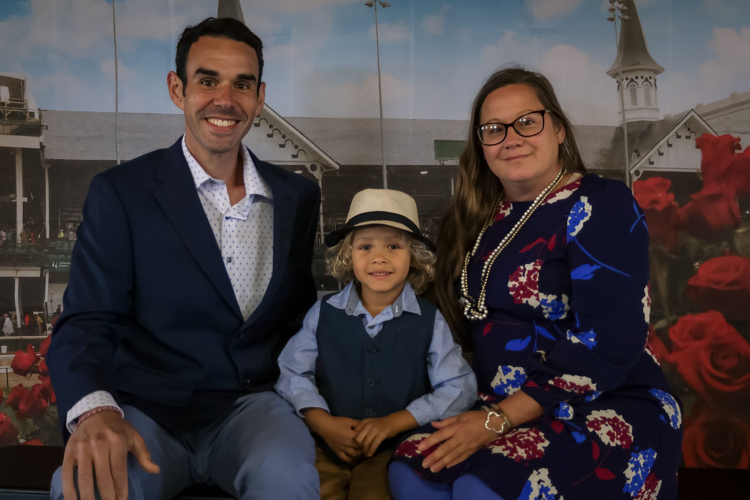 Family of three posing for a photo at Kentucky Derby, with roses backdrop, child wearing a hat.
