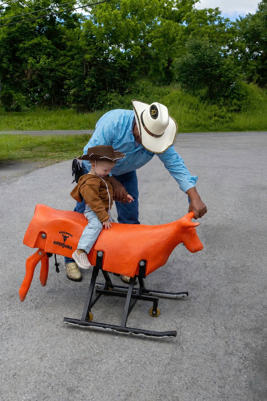 A man in a cowboy hat helps a young child sit on an orange mechanical bull toy outdoors near green trees.