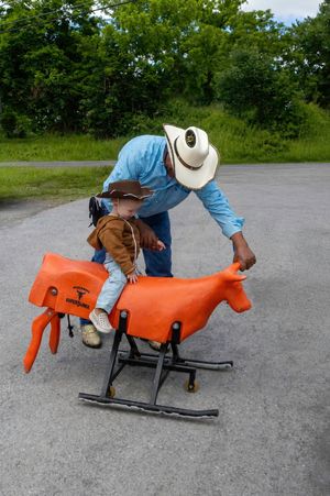 A man in a cowboy hat helps a young child sit on an orange mechanical bull toy outdoors near green trees.