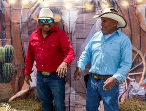 Two men in cowboy hats and western shirts pose in front of a rustic rodeo backdrop with cacti and hay.