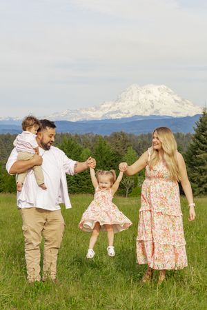 Family of four laughing and swinging toddler in green meadow with stunning Mount Rainier in background taken by yelm washington photographer