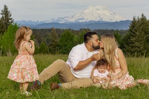 Family of four sitting in green field with parents kissing while two young kids play, mount rainier in back taken by washington photographer