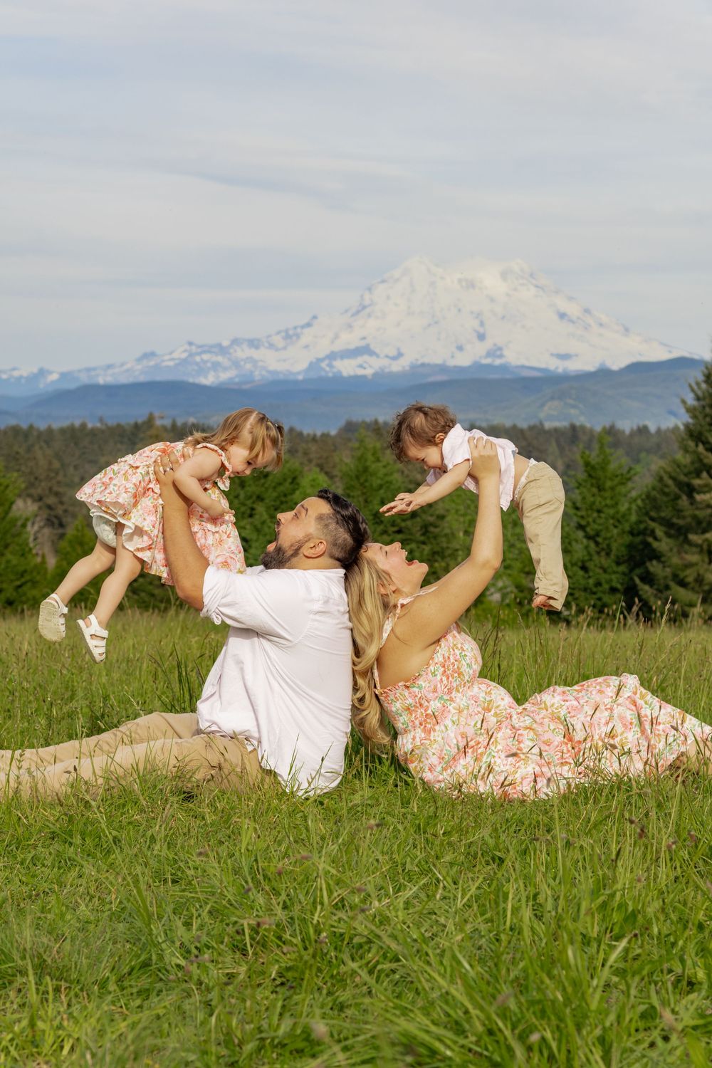 Family of four playing in green meadow, parents lifting two young kids with Mount Rainier behind them, captured by Roy WA photographer