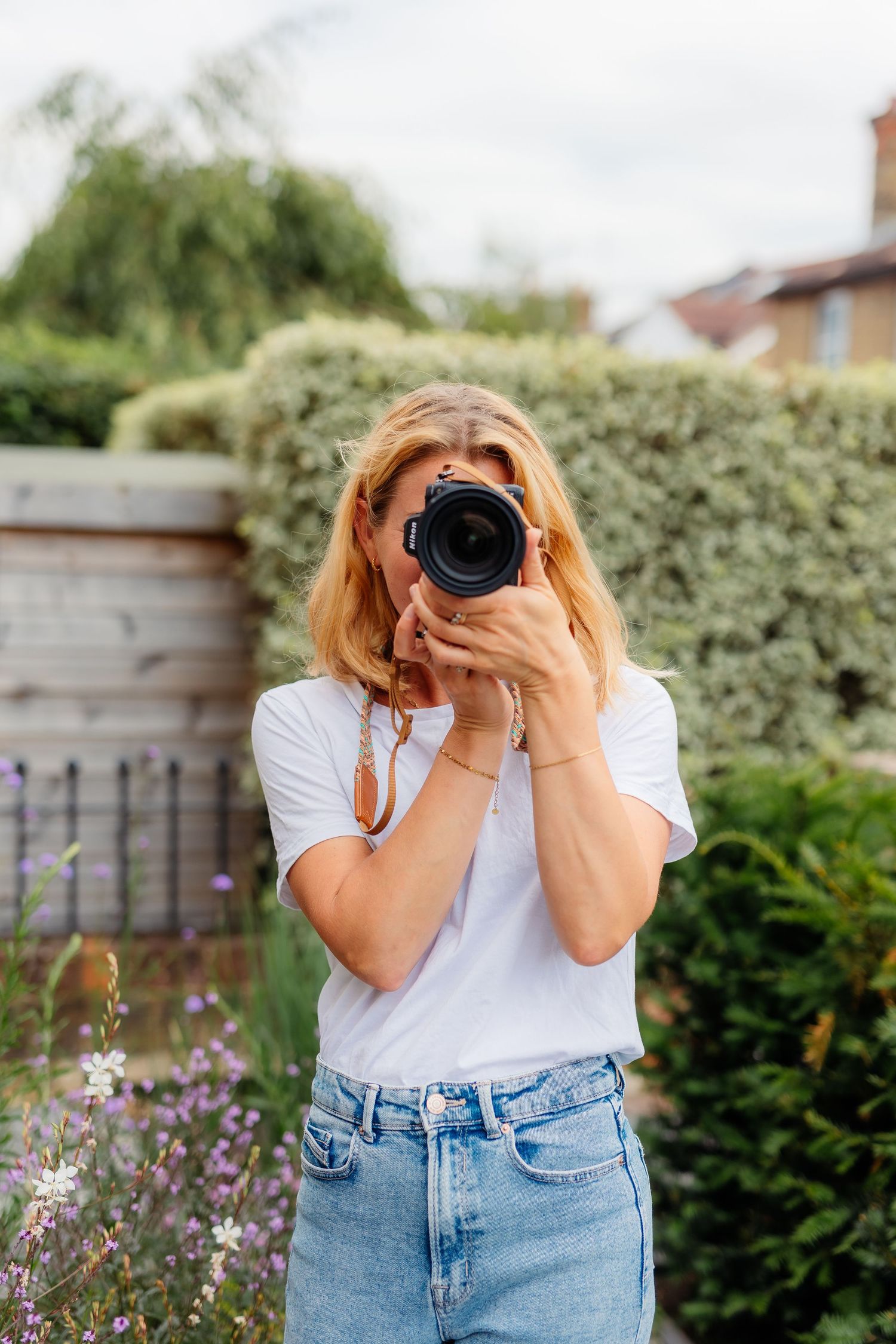 Blonde woman in white t-shirt and jeans photographing with a DSLR camera in a lush garden setting.
