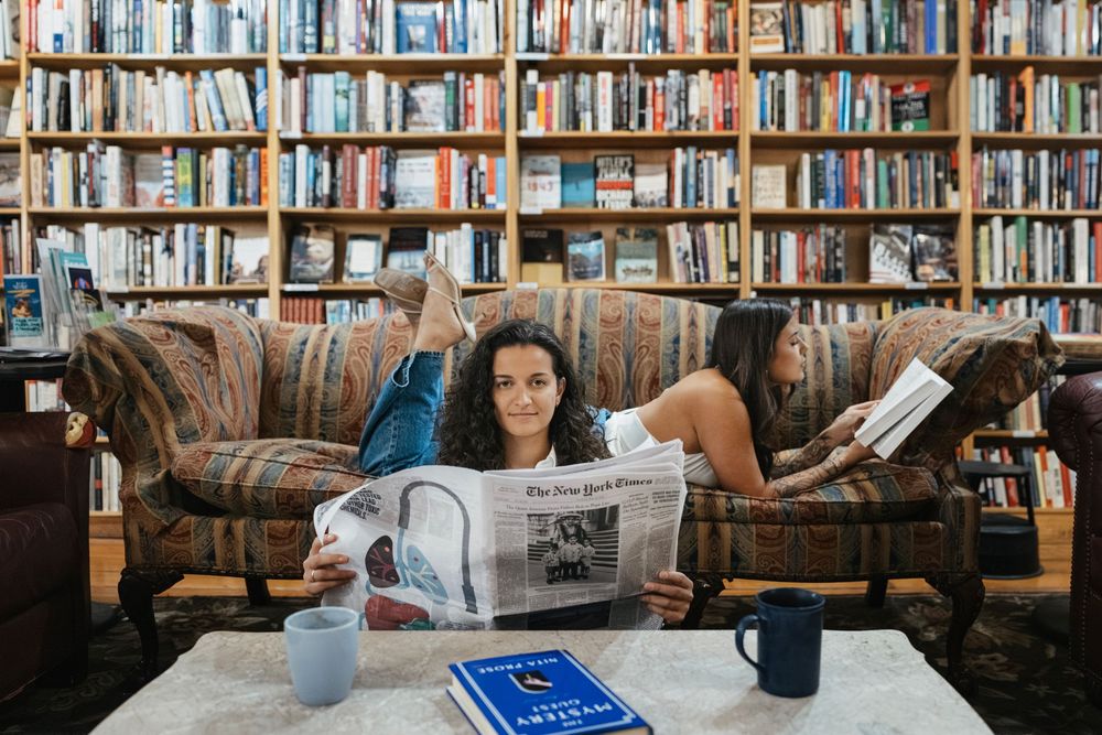 Woman relaxing on a colorful sofa reading the New York Times in a cozy home library filled with books.