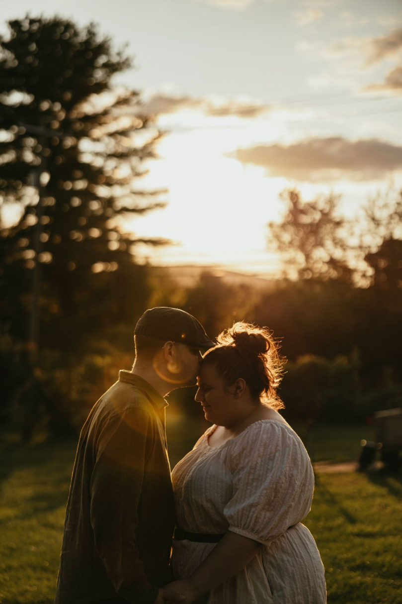 Couple embracing at golden sunset in a lush green park, backlit by warm glowing sky and trees.