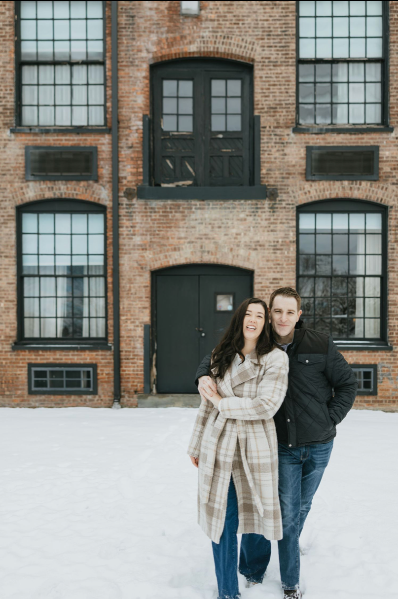 Couple smiling and embracing in snow outside a rustic brick building in winter, woman in plaid coat and man in black jacket.