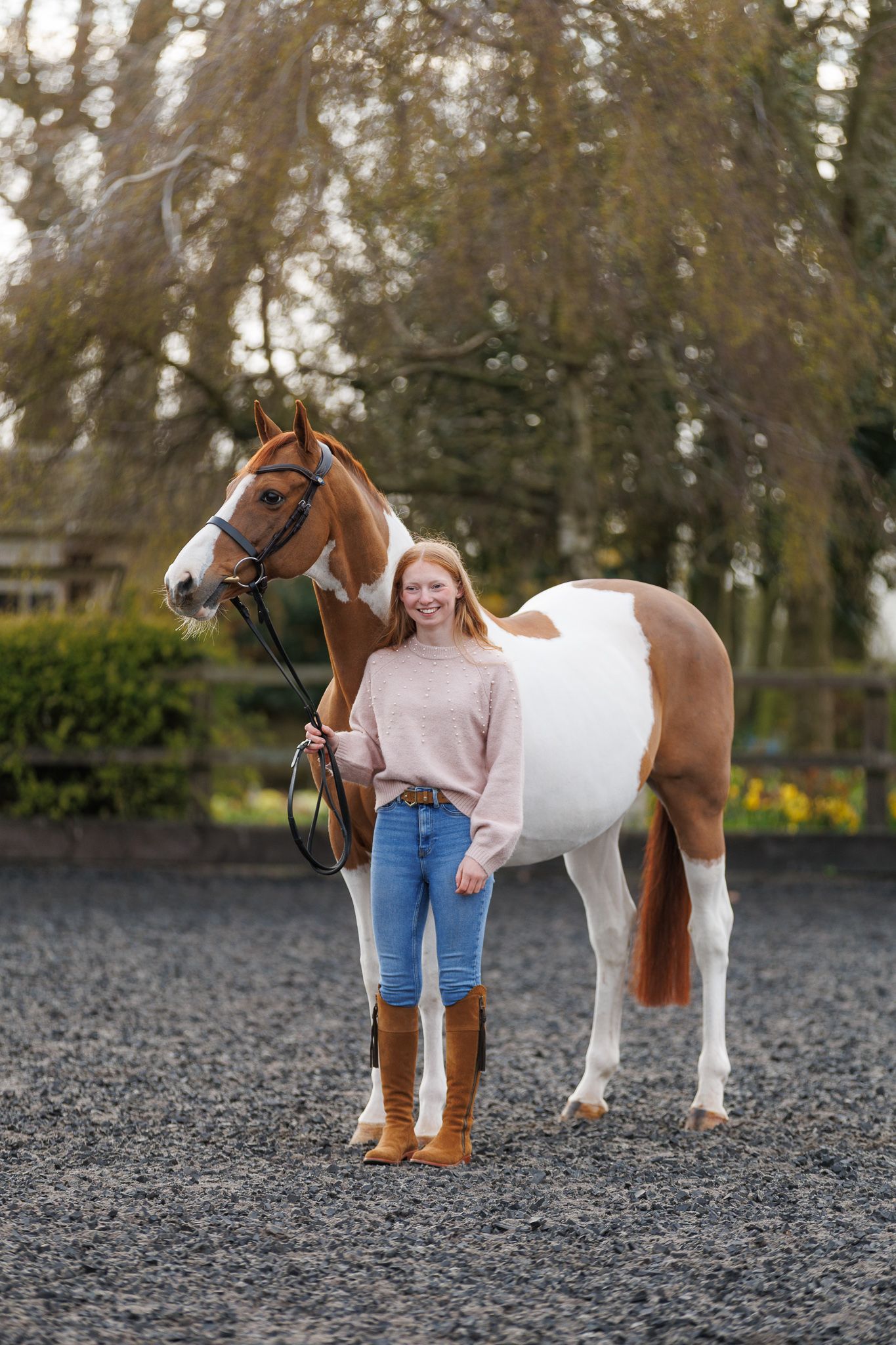 A beautiful bay horse wearing a black bridle stands next to someone in a white lace dress in an outdoor setting.