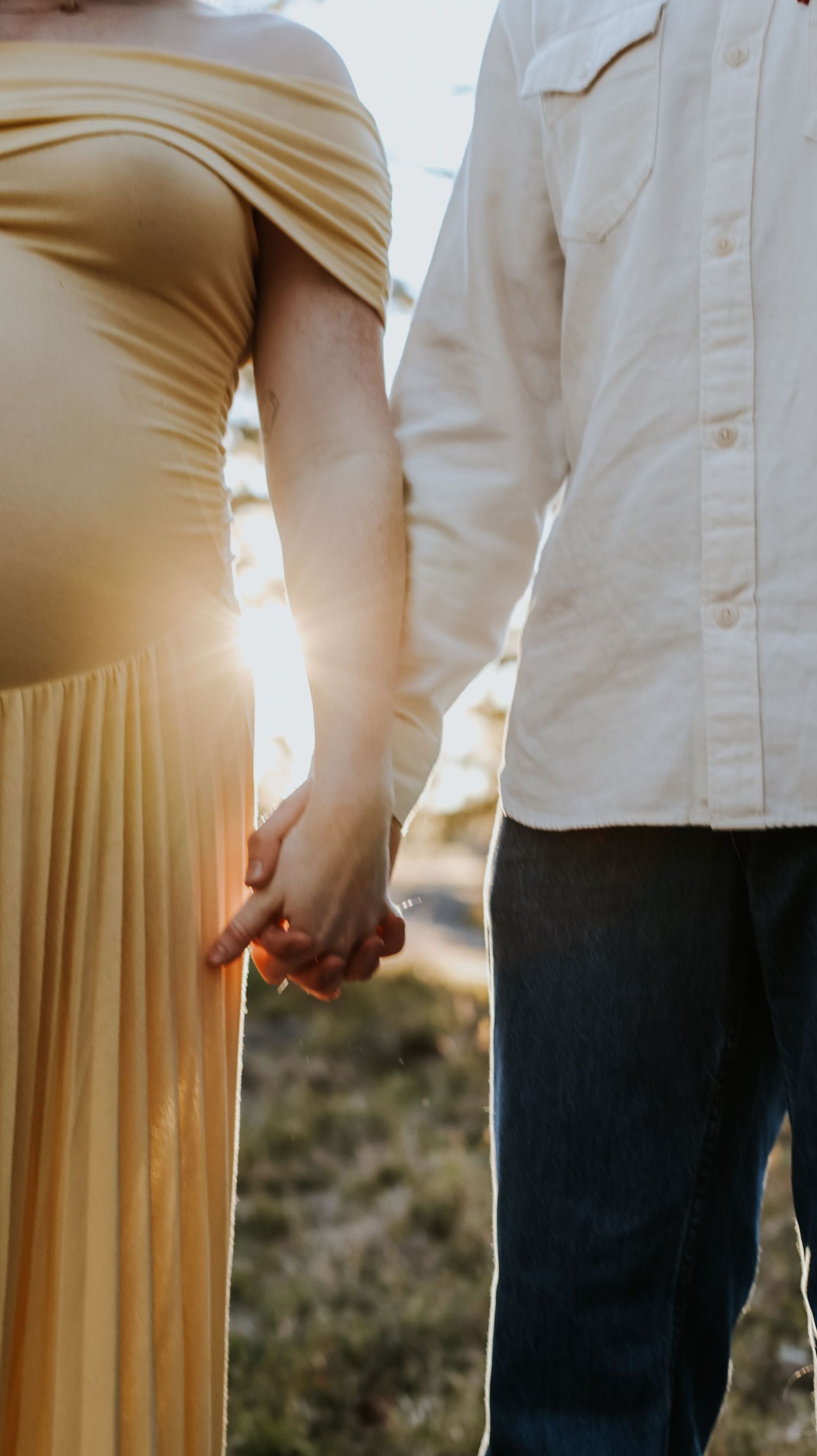 Pregnant couple holding hands at golden hour, woman in yellow off-shoulder gown, man in white shirt and jeans.