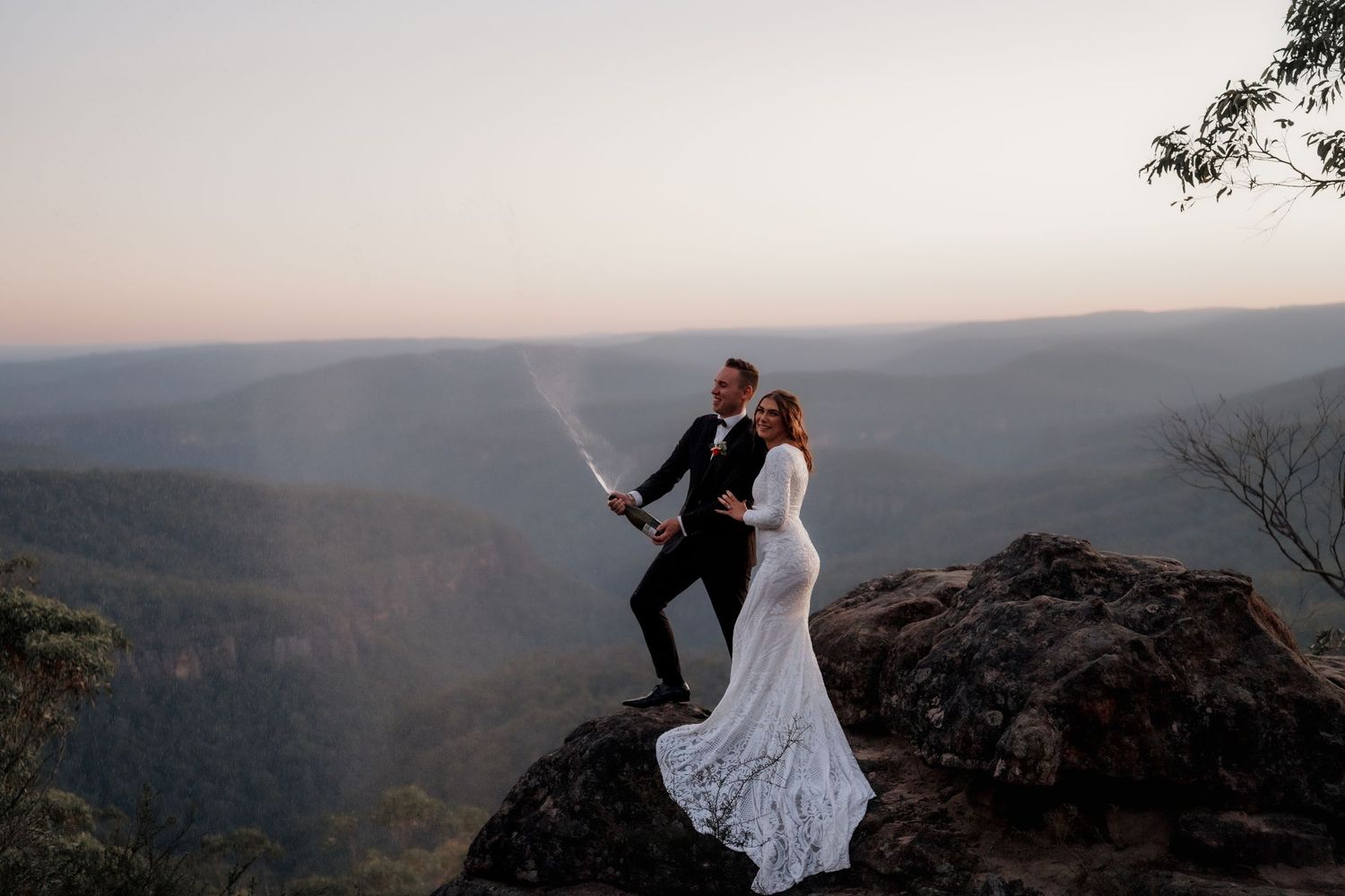 Bride and groom sabering champagne on a rocky cliff at sunset with stunning mountain valley views.