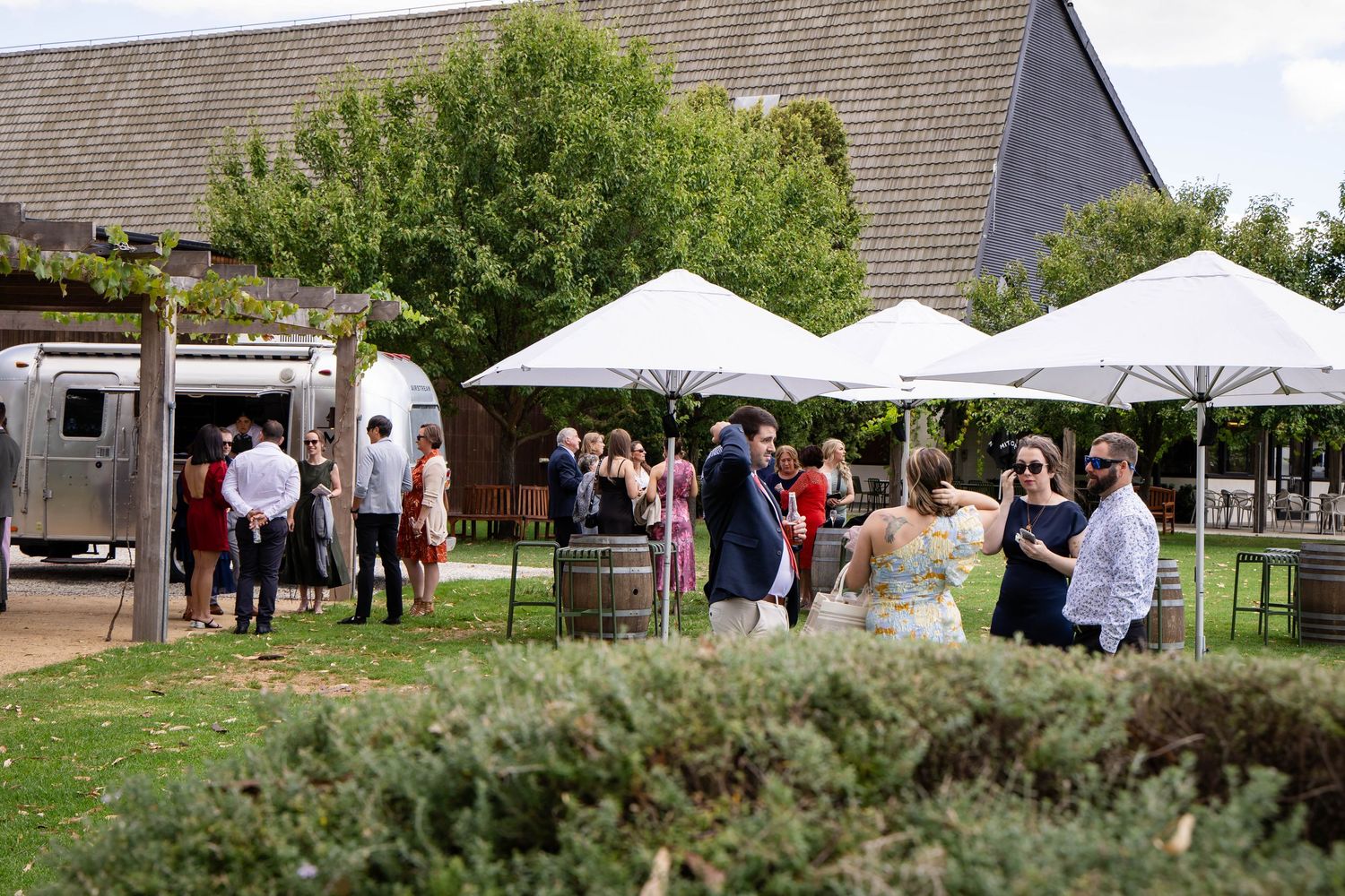Guests mingle outdoors at a winery event with white umbrellas, barrel decor, and an Airstream bar on a sunny day.