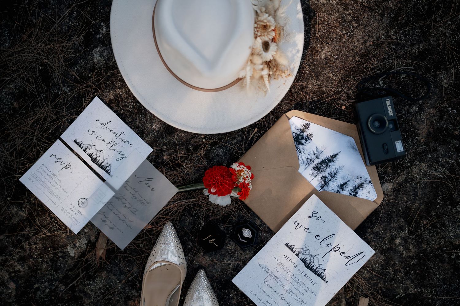 Rustic wedding stationery flatlay with white hat, vintage camera, red flowers, kraft envelopes, and elegant invitation suite on dark ground.