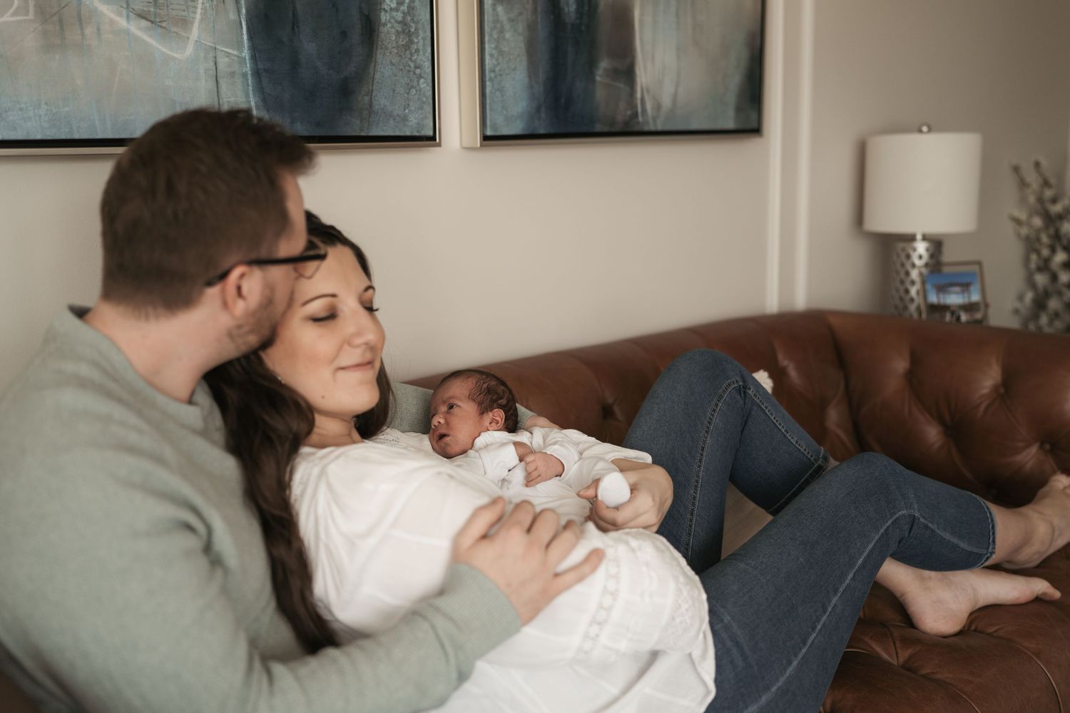 New parents sitting on brown leather sofa, mother holding newborn baby while father looks on lovingly in cozy living room.