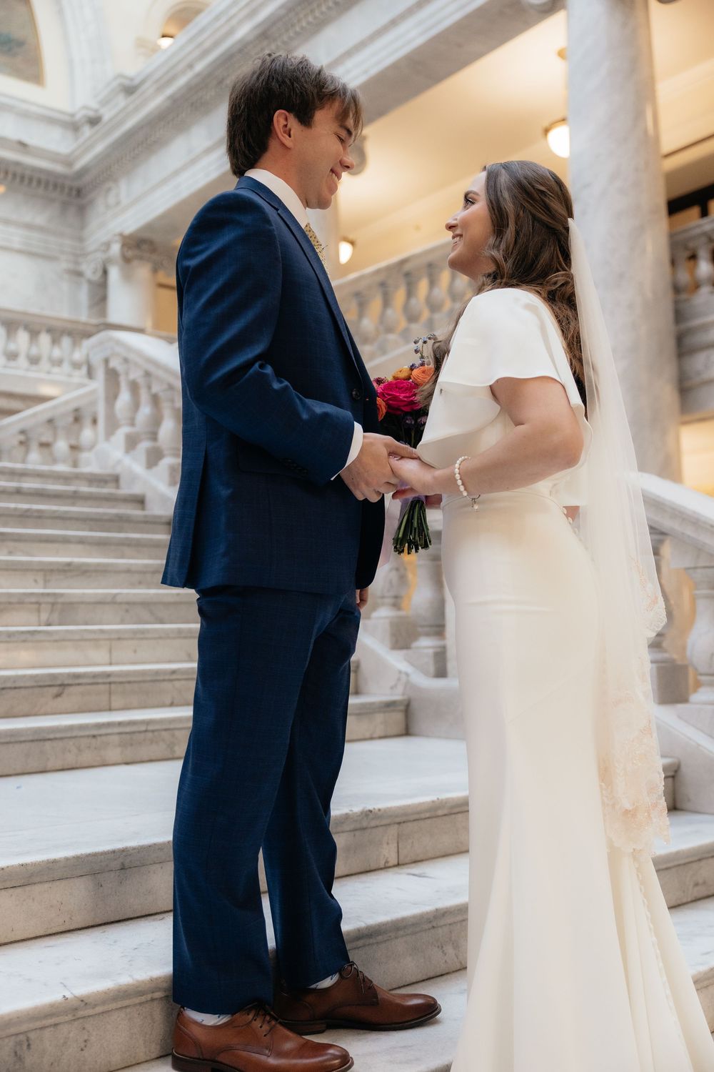 Bride and groom share emotional first look moment holding hands on marble staircase inside elegant capitol building.