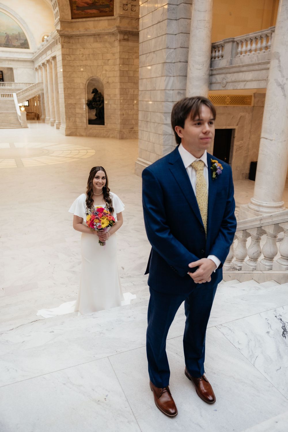 Groom in navy suit waits for first look as bride in white gown holds colorful bouquet inside marble capitol building.