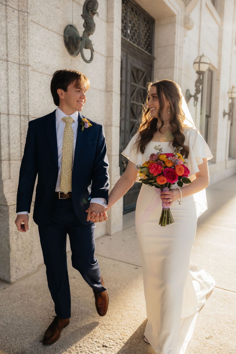 Happy newlyweds walking hand in hand outdoors, bride holding colorful bouquet in white gown.