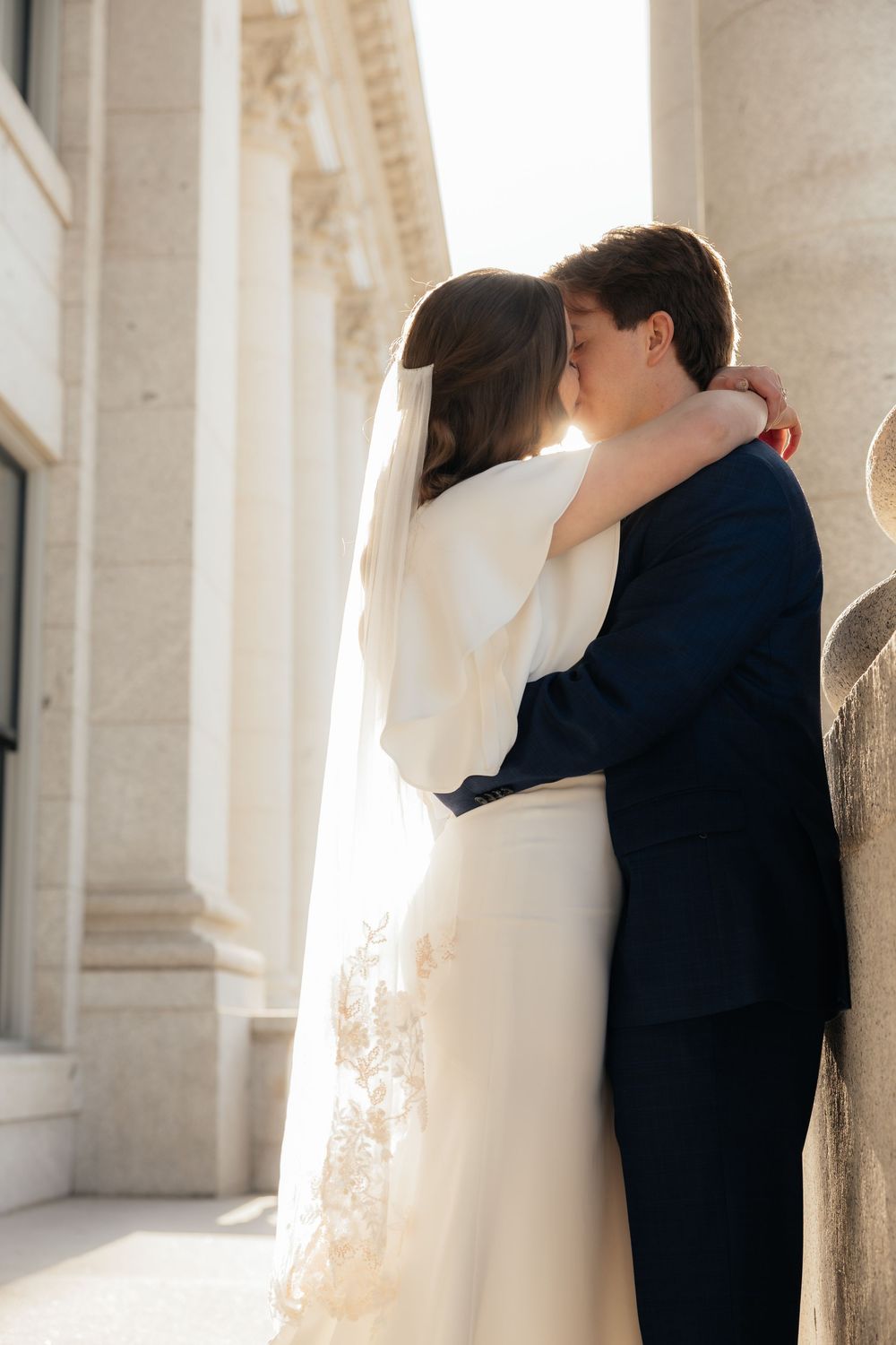 Bride and groom sharing a romantic kiss against sunlit stone columns on their wedding day.